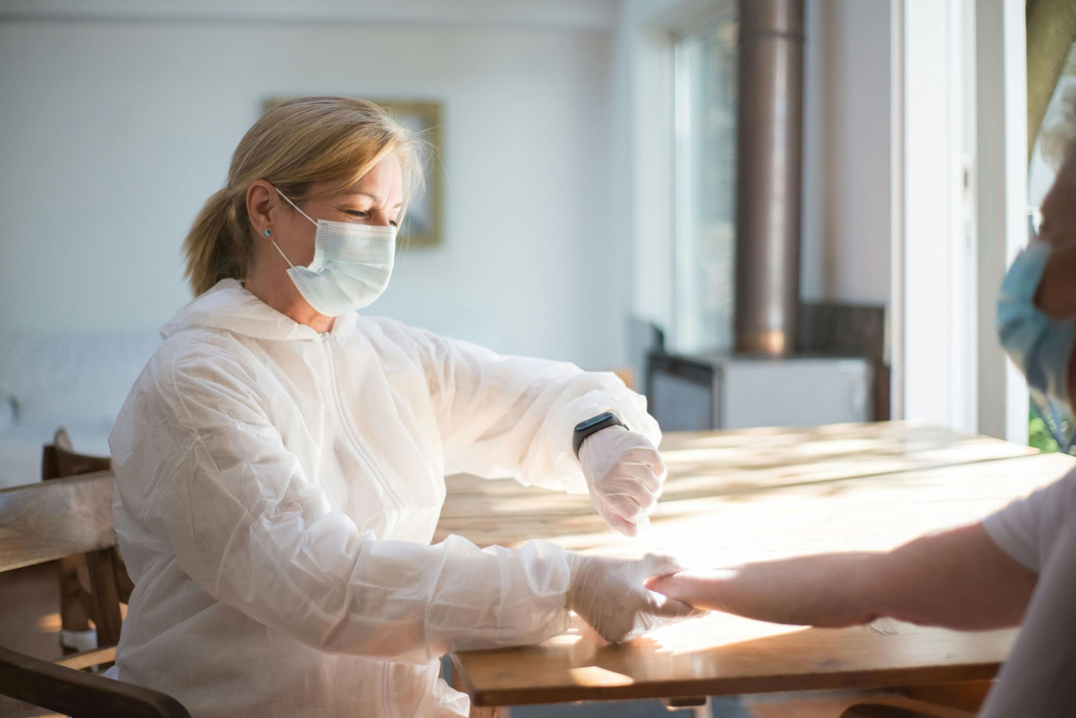 Healthcare worker in protective clothing providing care to a person at a wooden table in a bright room.