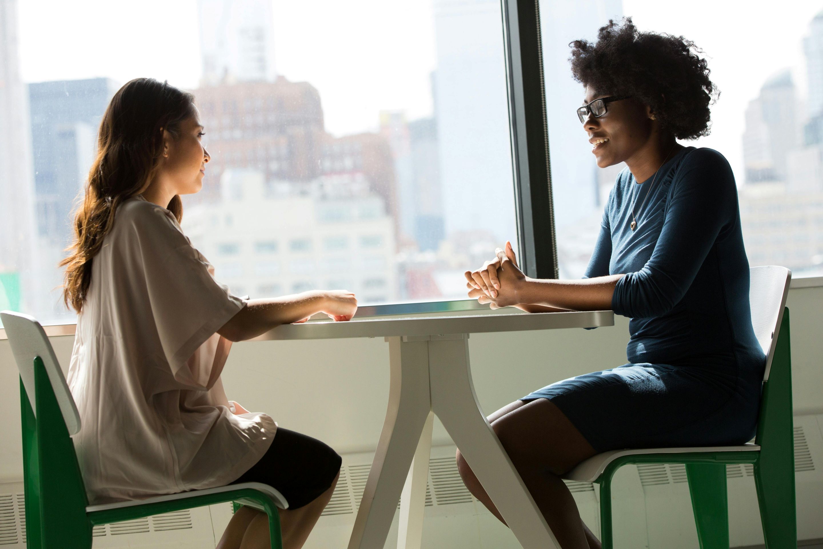 Two people sitting at a table in front of a large window, engaged in a conversation.