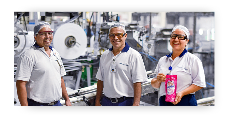 Dos hombres y una mujer sonriendo, parados frente a los equipos de la fábrica; la mujer sostiene un producto Suavitel