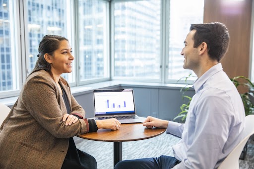 Homem e mulher sentados em uma mesa conversando. Há um notebook entre eles apresentado gráficos.