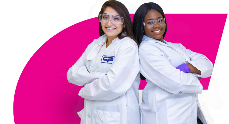 Две студентки в белых лабораторных халатах стоят спиной друг к другу в лаборатории, при этом улыбаясь