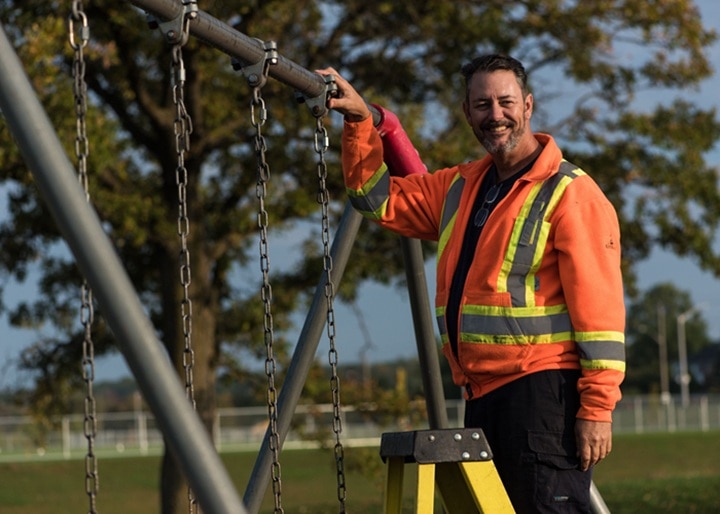 City of Kitchener employee smiles at the camera beside a swing set in a park.