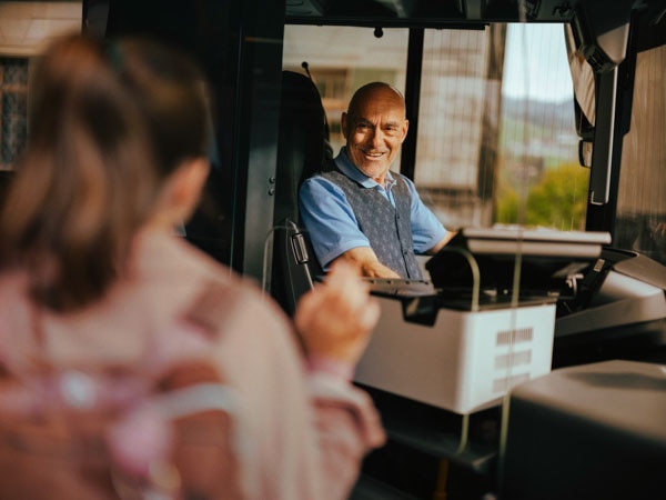 Homme en uniforme au volant d’un PostAuto saluant une passagère