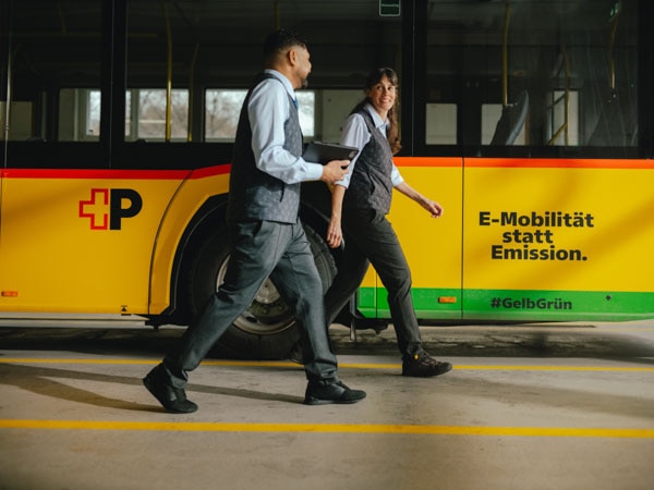 Frau und Mann in Uniform laufen an der Seite eines PostAuto entlang