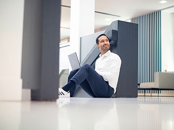 Dipendente di Allianz Suisee seduto comodamente sul pavimento con il computer portatile sulle gambe piegate.