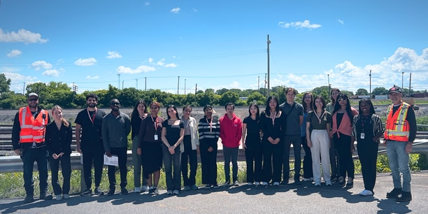 Group of employees gathered outdoors at a rail site with tracks and equipment in the background.