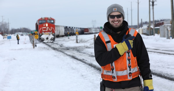Rail worker wearing a high-visibility vest, winter hat, gloves, and safety glasses standing beside snowy railway tracks, giving a thumbs-up, with a red CN locomotive and freight cars visible in the background.