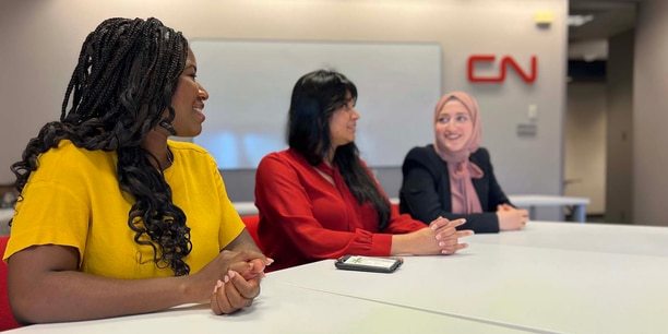 Three employees sit at a conference table in a modern office, engaged in discussion. A red CN logo is visible on the wall behind them, with a bright, collaborative workspace setting.