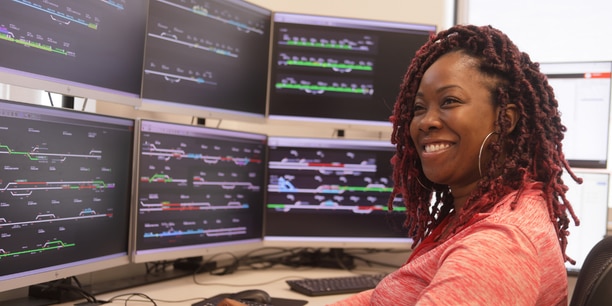 Smiling employee working at a control station with multiple monitors displaying rail system data.