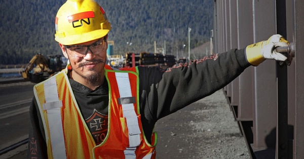 Rail worker wearing a yellow hard hat, safety glasses, and a high-visibility vest standing beside a railcar, with rail equipment and a mountainous landscape visible in the background.