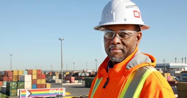 Rail worker wearing a white hard hat, safety glasses, and a high-visibility jacket standing in a rail yard, with stacked shipping containers and CN-branded equipment visible in the background.