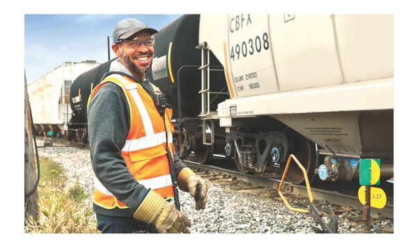 Travailleur ferroviaire portant un gilet de sécurité haute visibilité, des lunettes de protection et des gants, debout à côté d’un train de marchandises sur les rails, avec des wagons-citernes et des wagons visibles en arrière-plan.