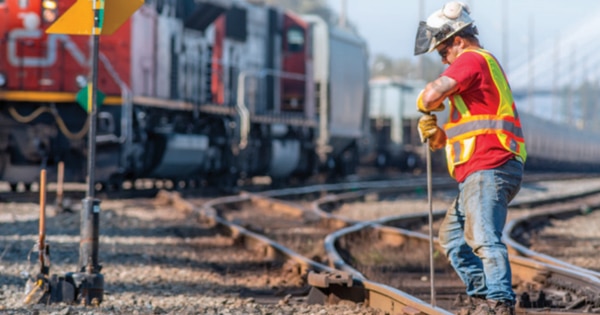 Rail worker wearing a hard hat, high-visibility vest, and gloves standing beside railway tracks, holding a tool while working near a track switch, with a CN locomotive and freight cars blurred in the background.