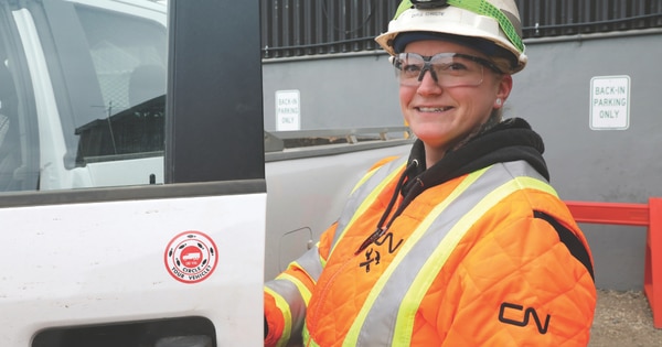 Rail worker wearing a hard hat, safety glasses, and a high-visibility jacket standing beside a white utility truck, with a CN logo visible on the sleeve and equipment in the background.