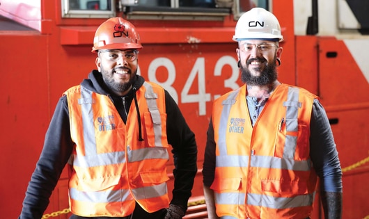 Deux travailleurs ferroviaires portant des gilets de sécurité haute visibilité et des casques se tiennent devant une locomotive rouge de CN