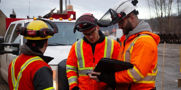 Three workers wearing helmets, safety glasses, and high-visibility jackets stand beside a utility truck. 