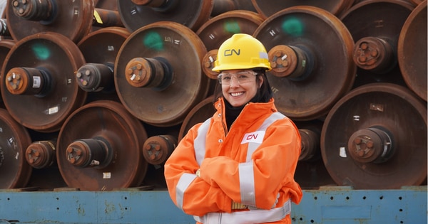 Rail worker wearing a yellow hard hat, safety glasses, and a high-visibility jacket standing with arms crossed in front of stacked train wheels, with industrial equipment visible in the background.