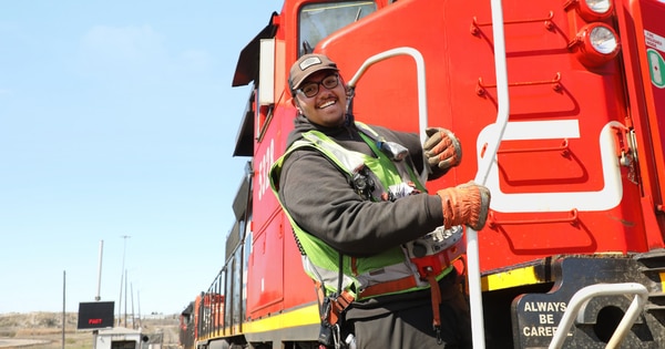 Rail worker wearing a high-visibility vest, gloves, and a cap standing on the steps of a red CN locomotive, holding a handrail with rail cars visible in the background under a clear sky.