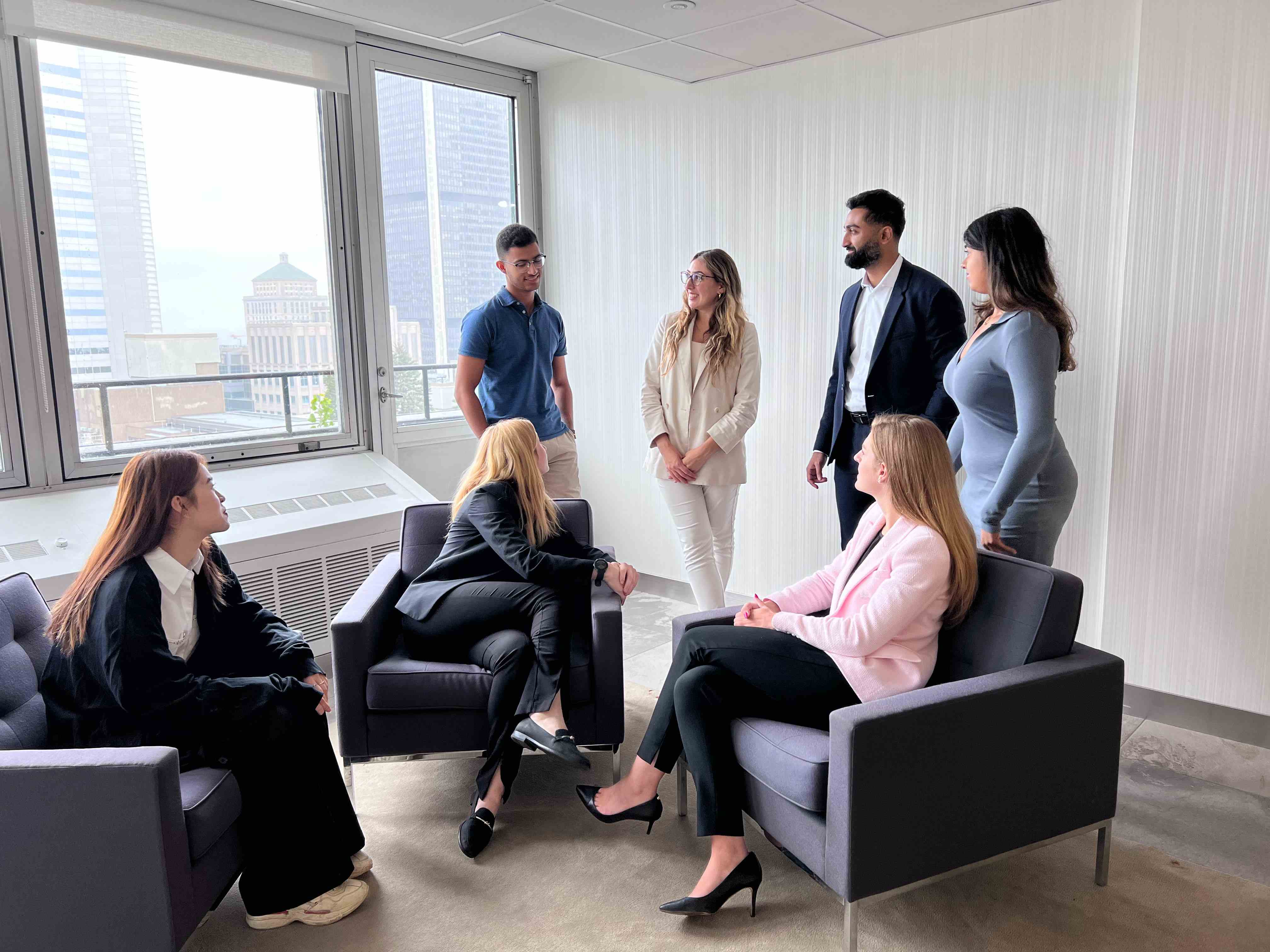 Seven employees in business attire are gathered in a bright office space with large windows overlooking a city. Some are seated in lounge chairs while others stand, engaged in a group discussion.