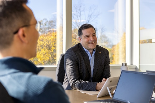 An image of two men seated at a table with laptops in a modern office setting.