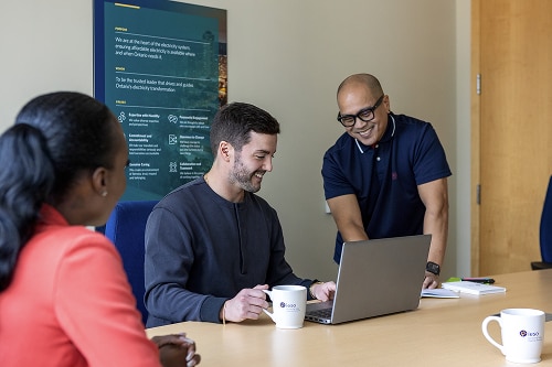 An image of two men smiling over a laptop, with a third person watching.