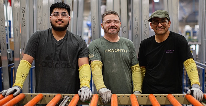 Tres trabajadores de una fábrica que llevan lentes de seguridad y protectores amarillos para los brazos están uno al lado del otro, sonriendo. Están en un entorno industrial con vigas metálicas.