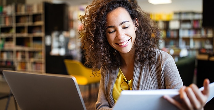 Mujer sonriente con el pelo rizado en una biblioteca mira un documento, sentada junto a un portátil. La escena transmite concentración y satisfacción. Estanterías de libros al fondo.