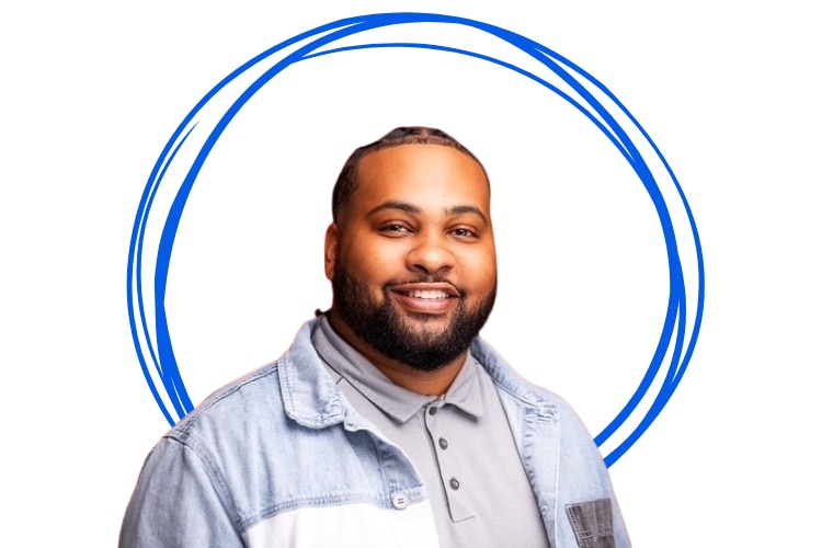A man sporting a beard and a denim jacket, posing casually against a plain background.