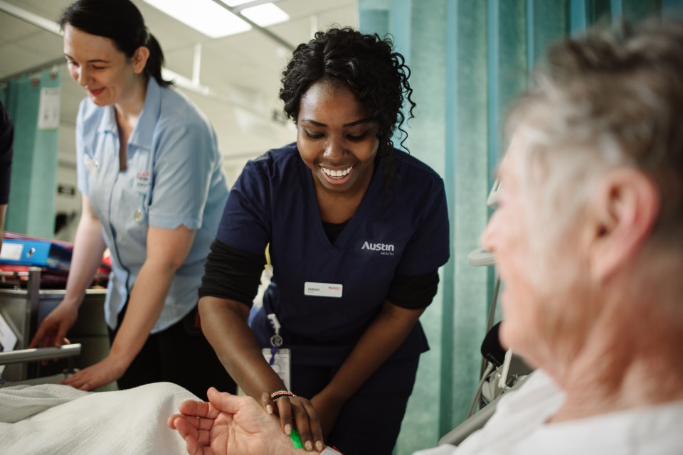 Two nurses in scrubs sit at a nurse station in a busy ward looking at a computer