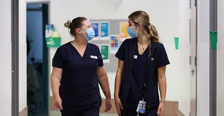 Two Austin Health clinical staff members wearing scrubs and face masks have a friendly chat while walking down a hospital corridor.